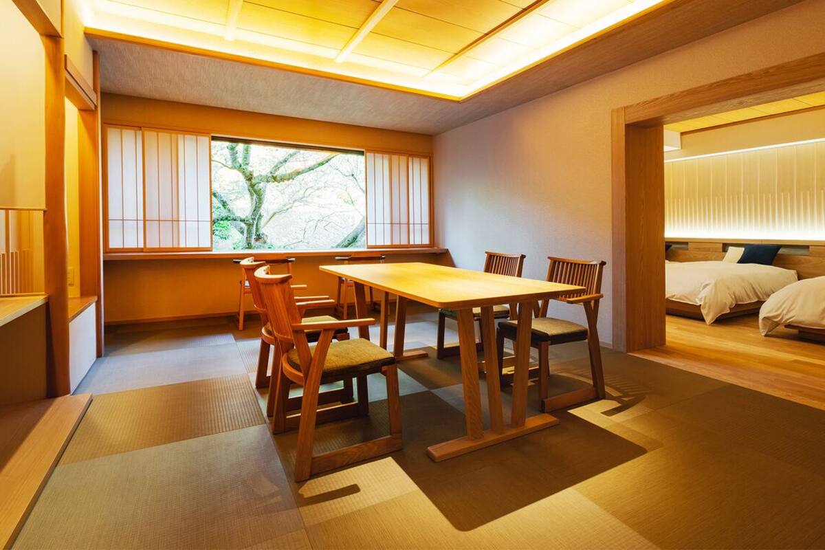 Una habitación tradicional japonesa con suelo de tatami, una mesa baja con sillas, una cama con cabecero de madera y vistas a la vegetación a través de una ventana.
