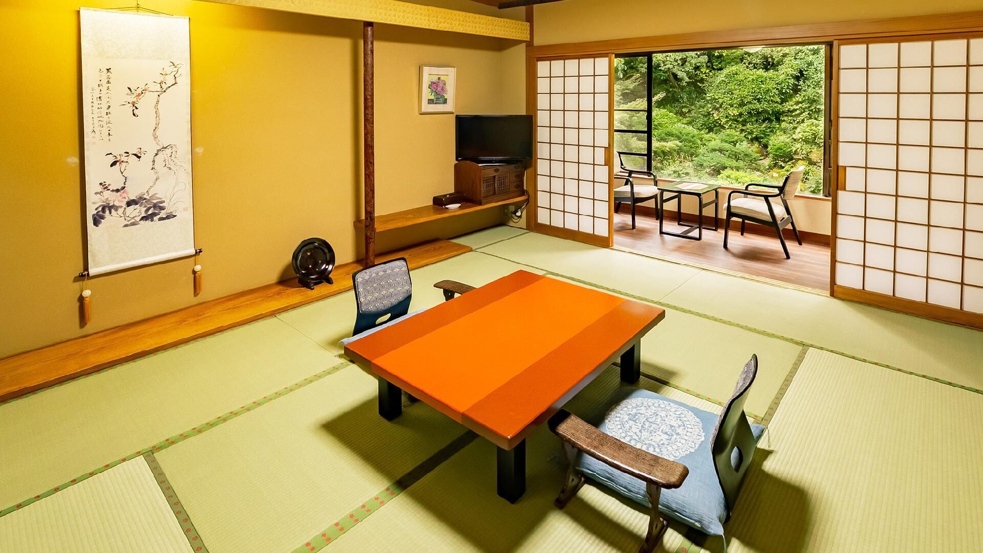 A tatami mat room with a low table and chairs, a television, and a view of greenery through shoji screens.