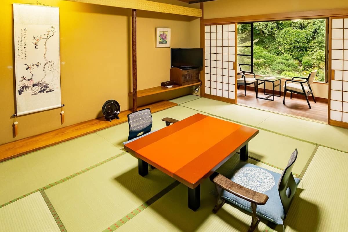 A tatami mat room with a low table and chairs, a television, and a view of greenery through shoji screens.