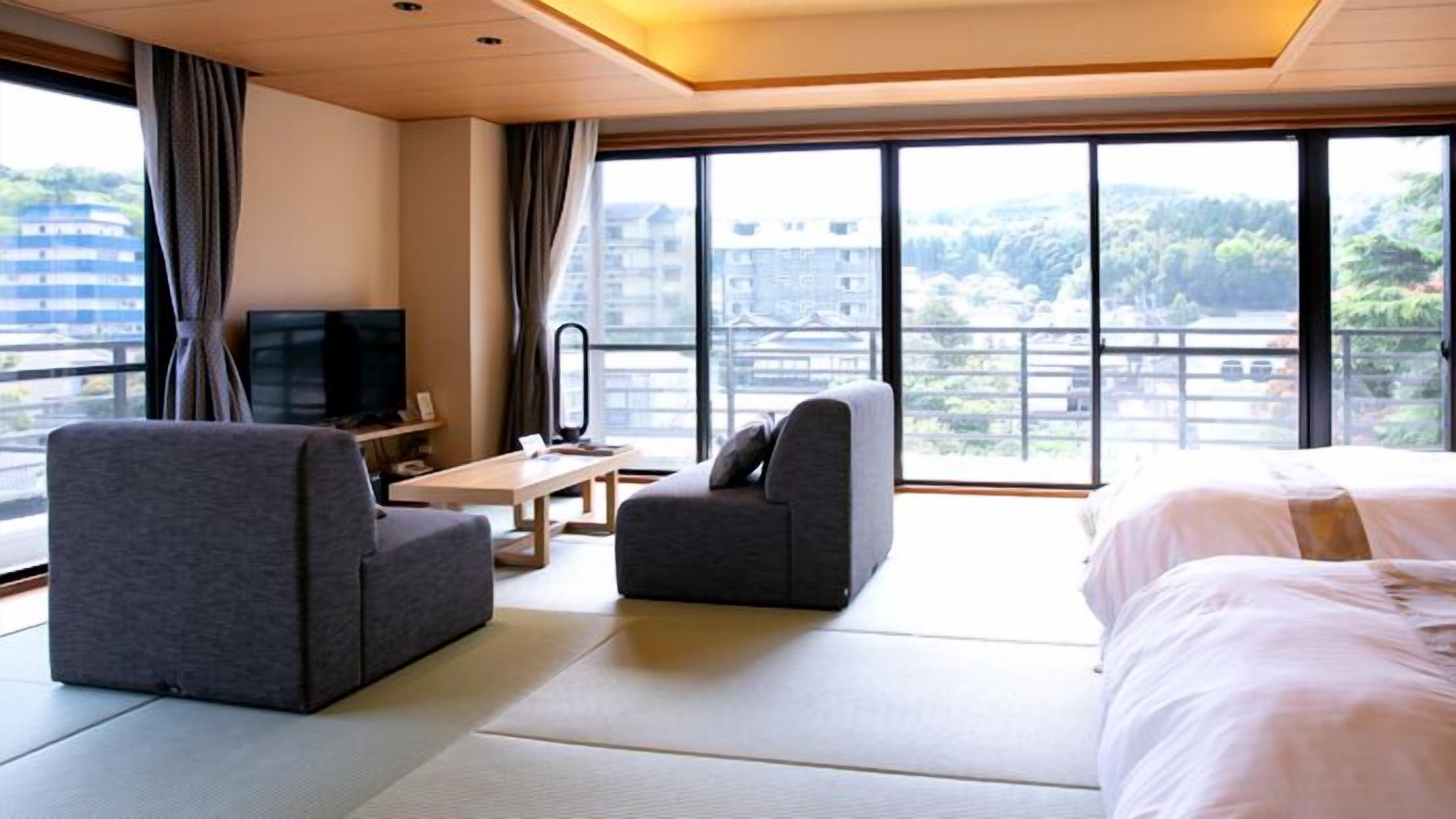 A hotel room with a bed, two armchairs, a TV, a desk, and large windows with a view of buildings and trees.