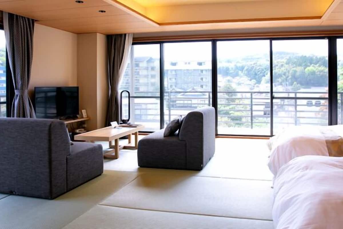 A hotel room with a bed, two armchairs, a TV, a desk, and large windows with a view of buildings and trees.