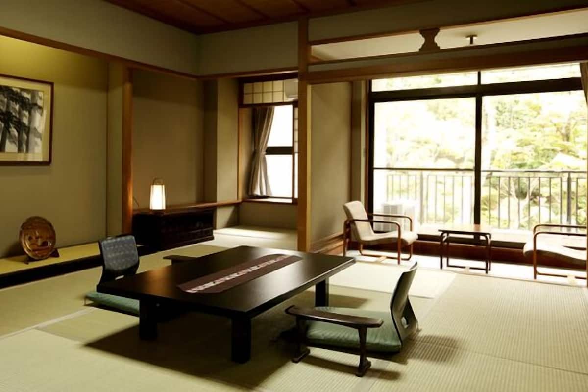 Una habitación japonesa tradicional con suelo de tatami, una mesa baja, sillas, una pintura y una ventana con vistas.