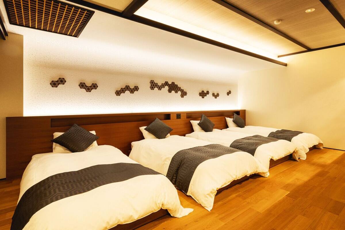 Habitación de hotel con tres camas, cabecero de madera, lámpara de pared y rejilla de ventilación en el techo.