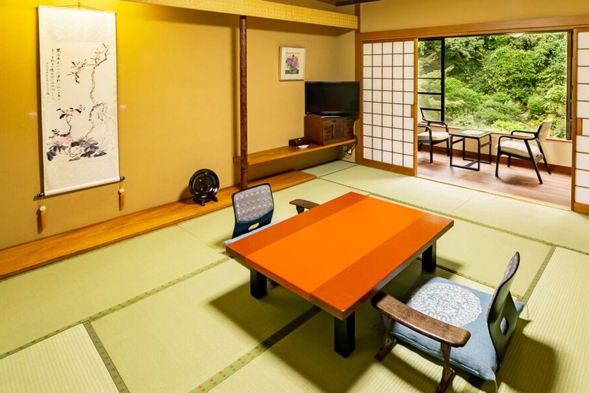 Una habitación con tatamis, una mesa baja, sillas, un televisor y una puerta corrediza que da a un balcón con vistas a la vegetación.