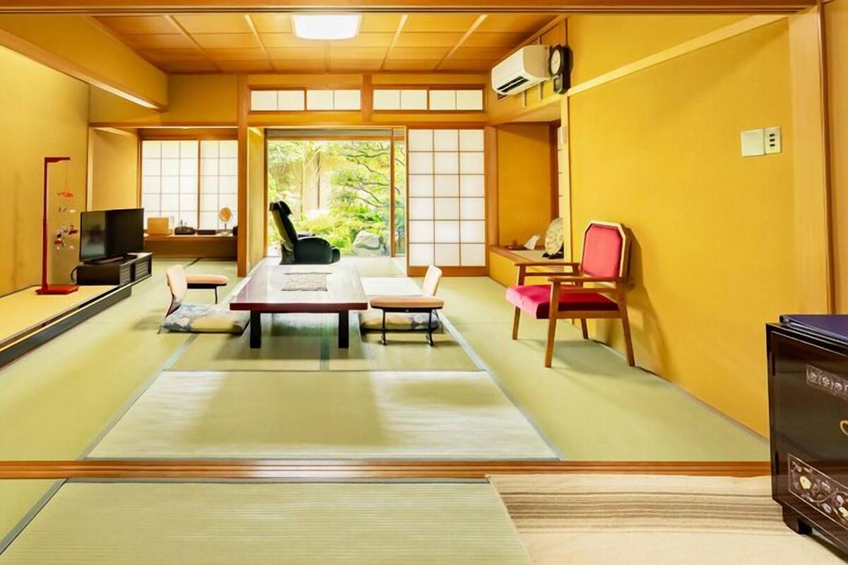 Una habitación japonesa tradicional con suelo de tatami, una mesa baja, sillas, un televisor y vistas al verdor.