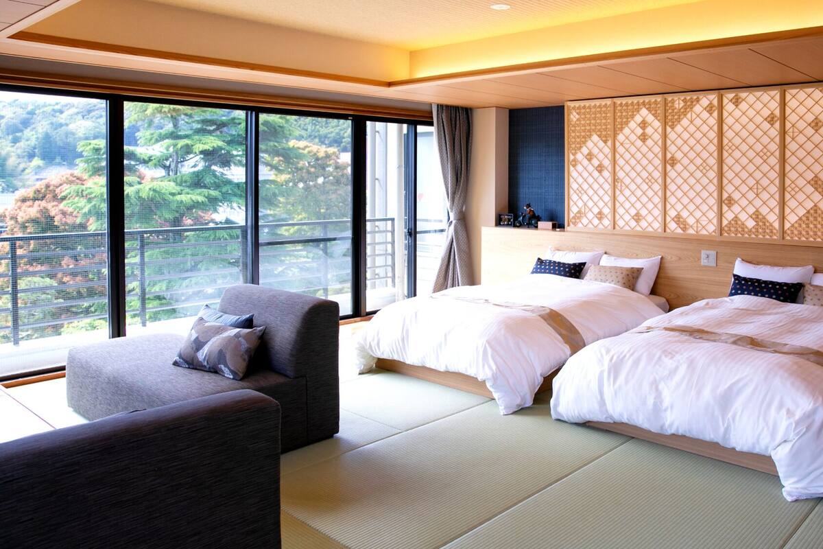 A hotel room with two beds, a sofa, tatami flooring, a large window with a view, and a decorative screen.