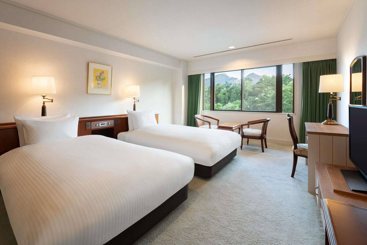 A hotel room with two beds, a desk with a chair, a TV, and a window with a view of greenery.