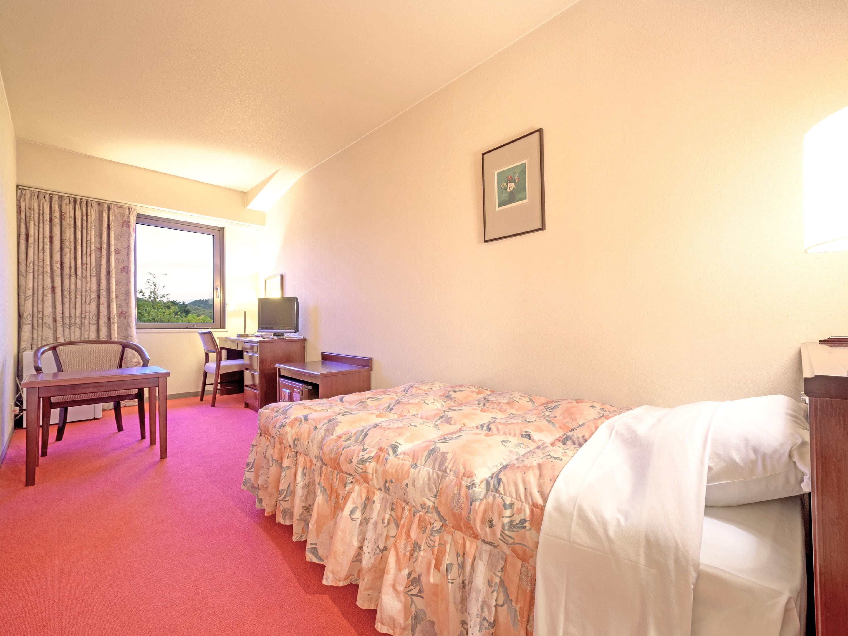 A hotel room with a bed, a desk with a computer, a chair, and a window with curtains.