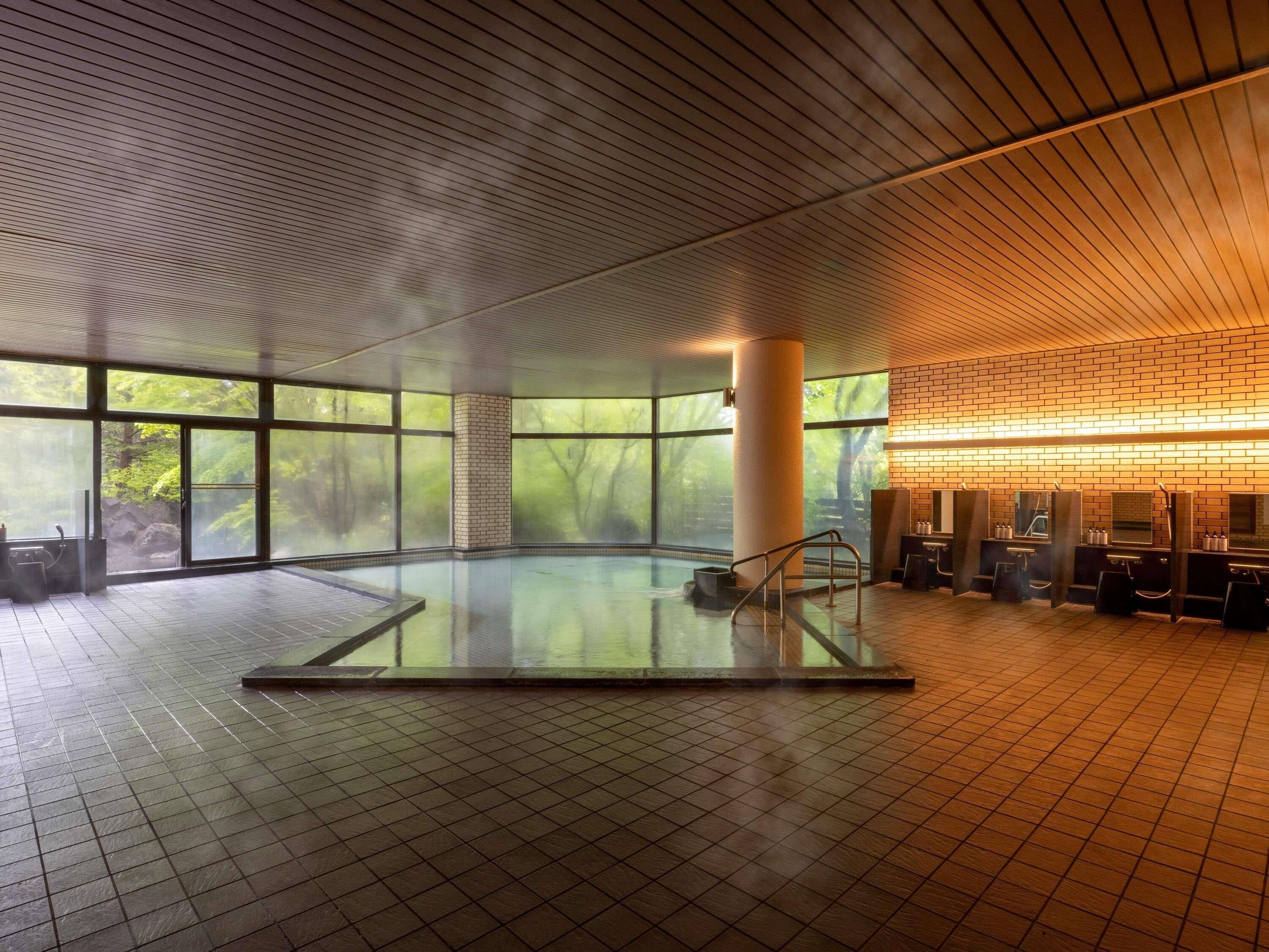 A large indoor swimming pool with a tiled floor, surrounded by large windows offering views of greenery.