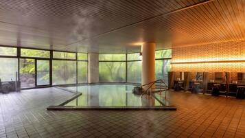 A large indoor swimming pool with a tiled floor, surrounded by large windows offering views of greenery.