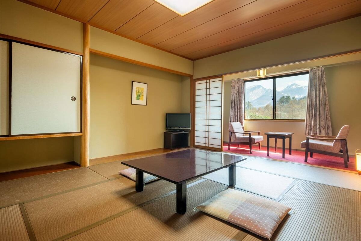 A traditional Japanese room with tatami flooring, a low table, a TV, and a view of mountains through a large window.
