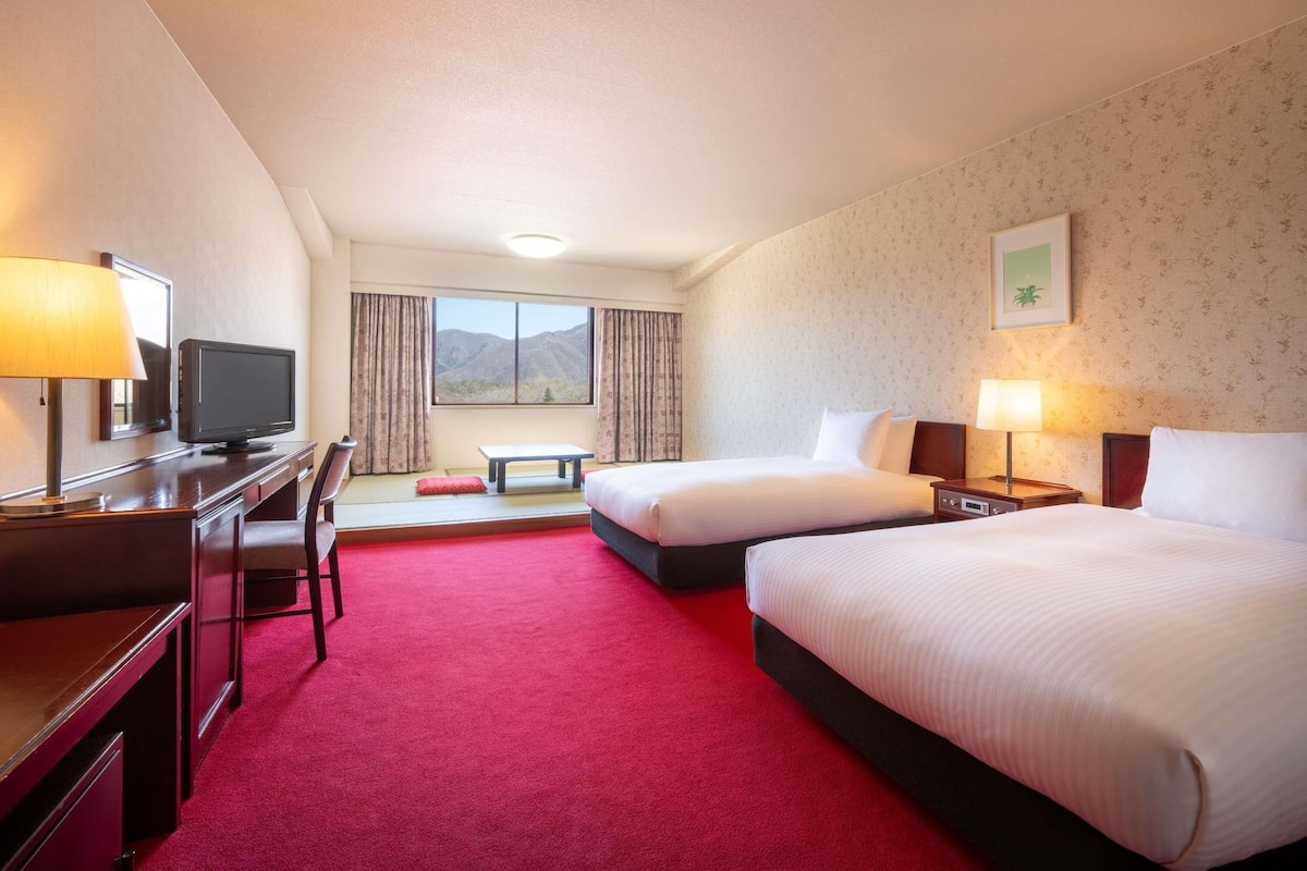 A hotel room with two beds, a desk with a TV, a window with a mountain view, and a red carpet.