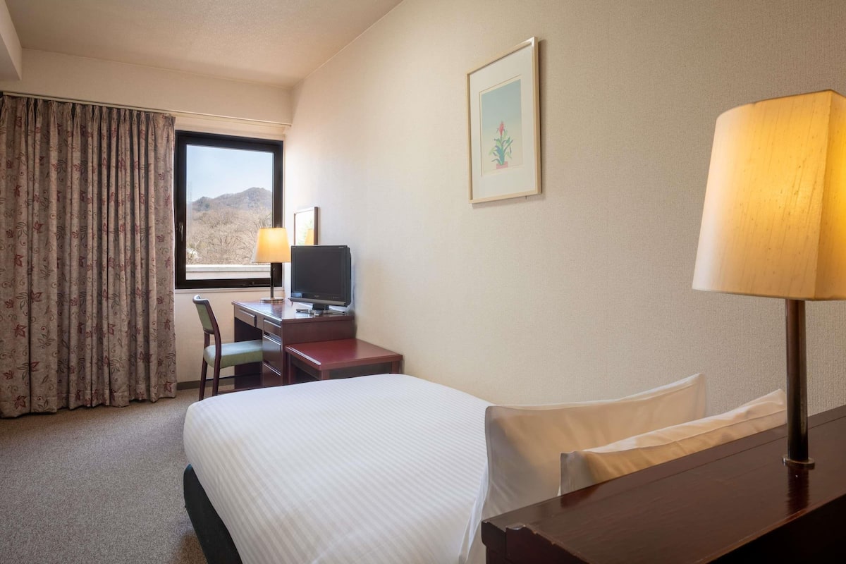 A hotel room with a bed, a desk with a computer, a chair, a window with a view of the mountains, and a lamp.