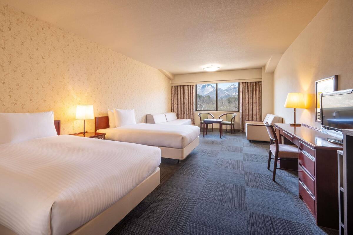 A hotel room with two beds, a desk, a chair, a TV, and a window with a mountain view.