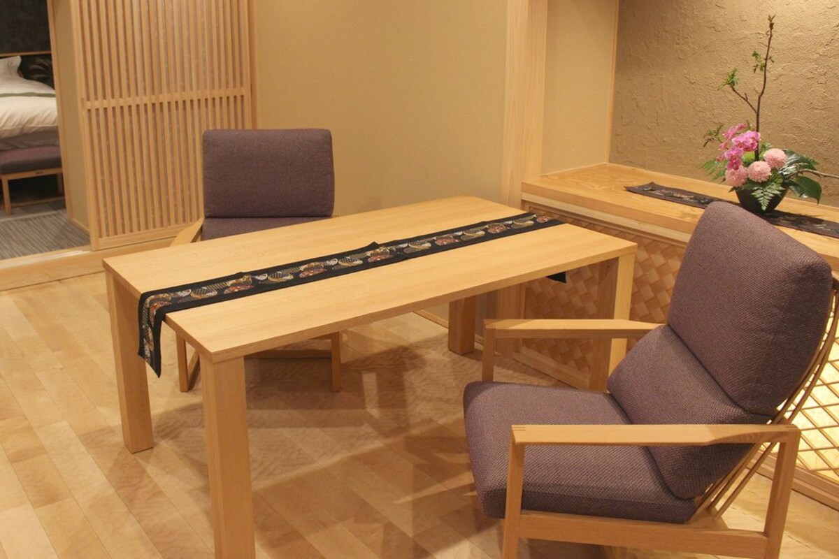 A room with a wooden table, two chairs, a bed with a headboard, a wooden partition, and a small table with a vase of flowers.
