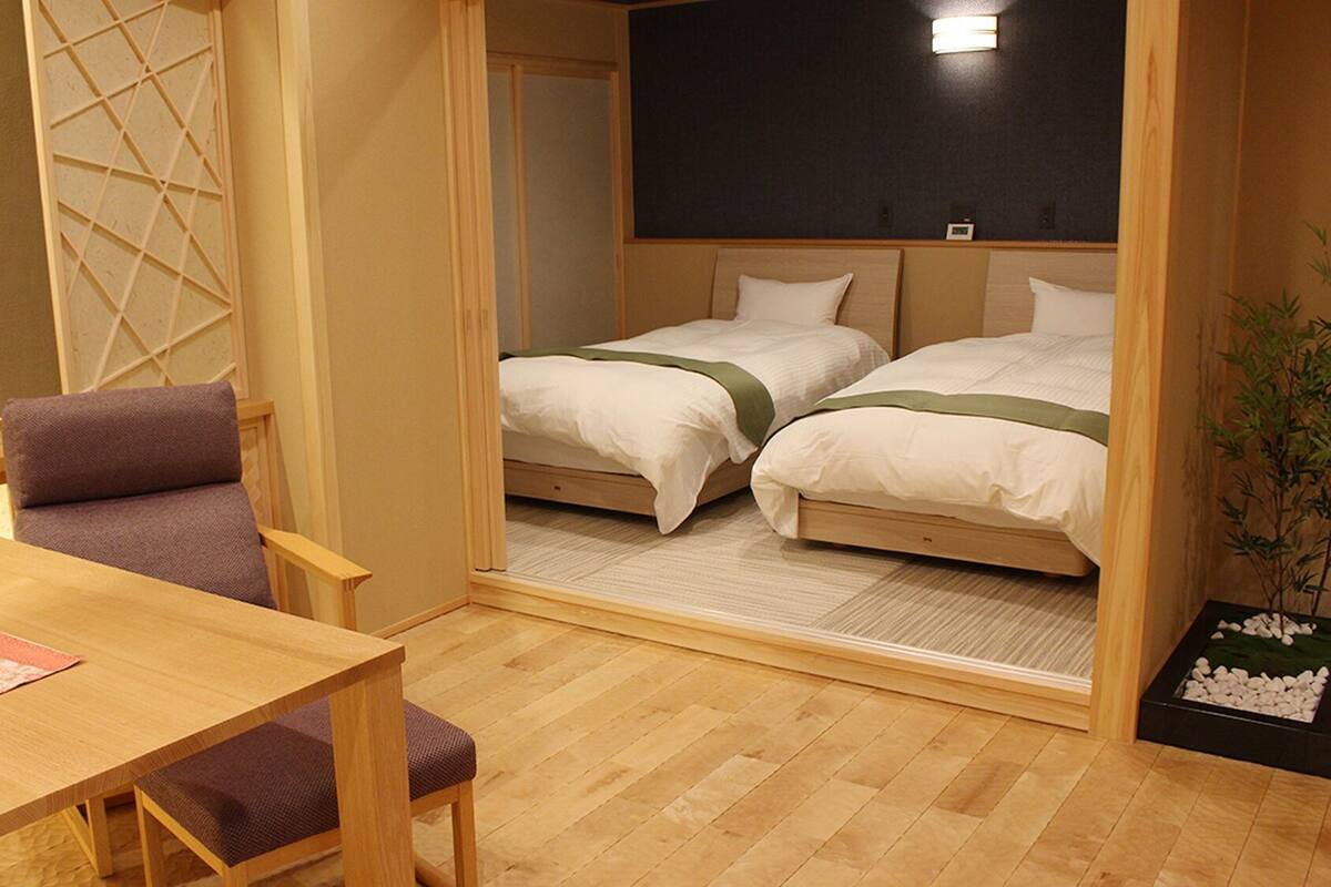 A room with a wooden table, a chair, two beds with white bedding, a small plant, and a blackboard on the wall.