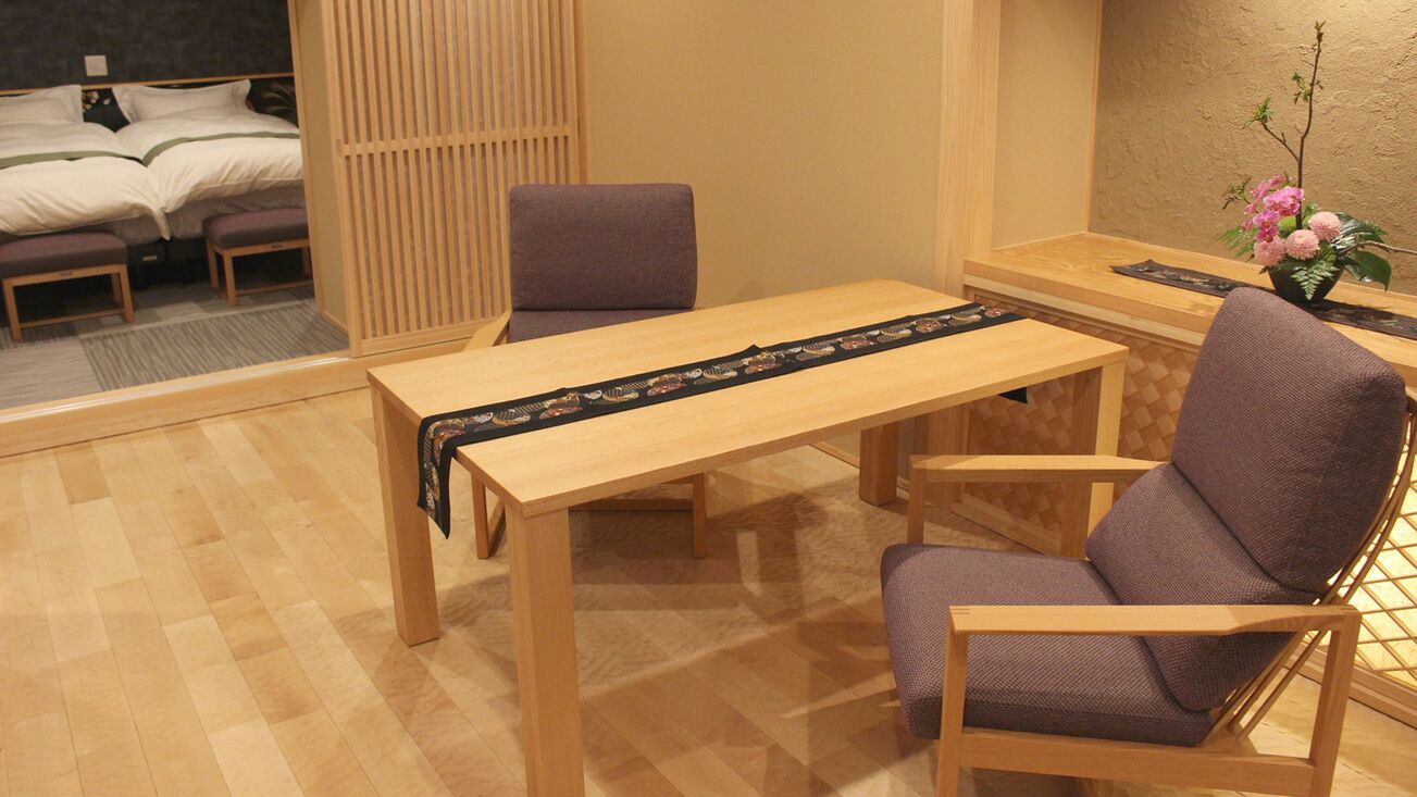 A room with a wooden table, two chairs, a bed with a headboard, a wooden partition, and a small table with a vase of flowers.