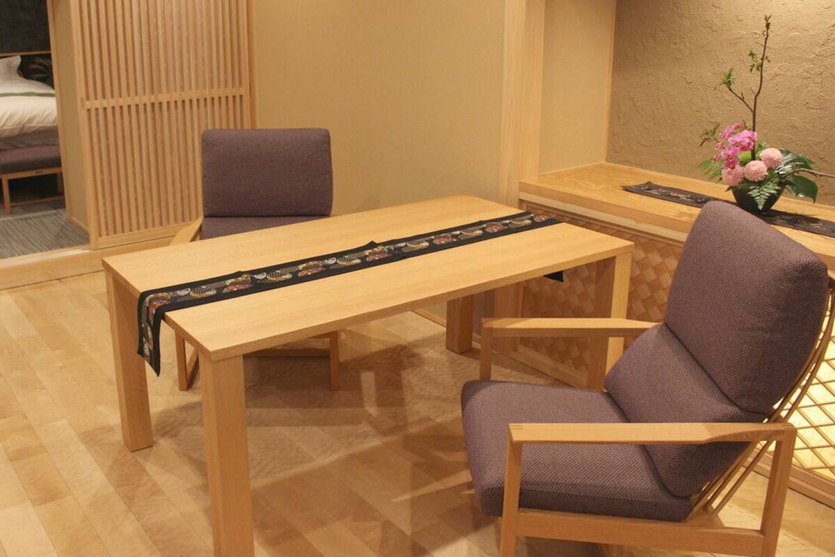 A room with a wooden table, two chairs, a bed with a headboard, a wooden partition, and a small table with a vase of flowers.