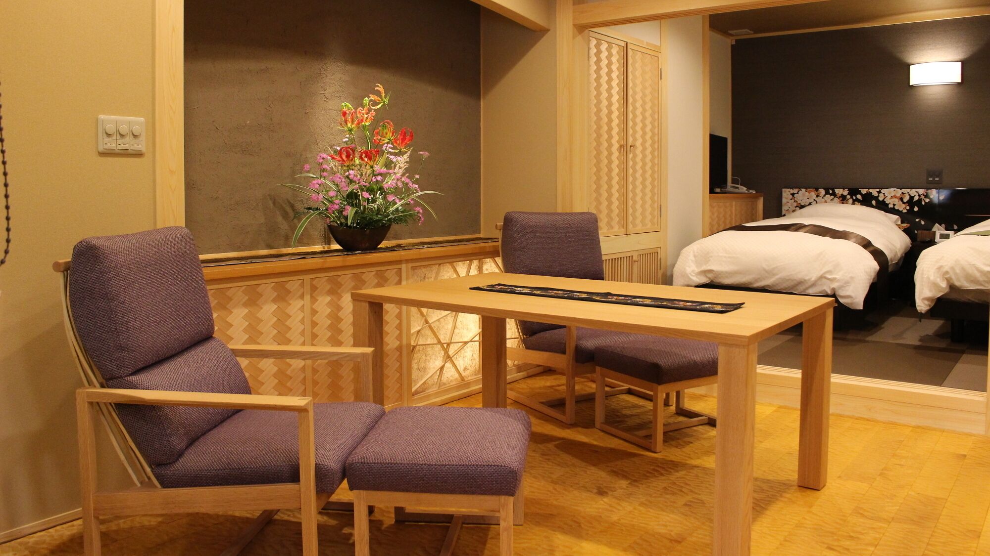 A hotel room with a wooden table, two purple chairs, a flower vase, and a bed with white bedding.