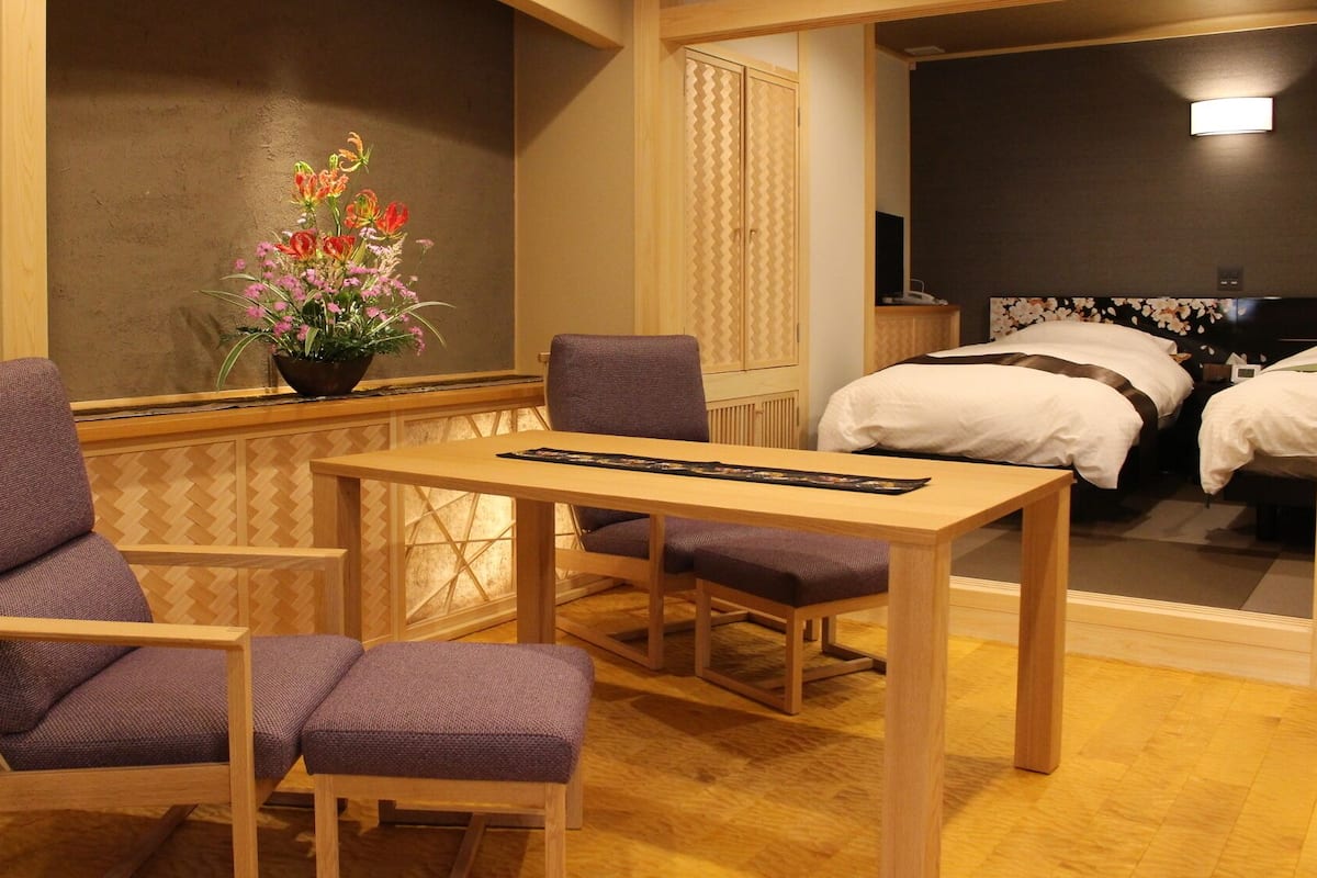 A hotel room with a wooden table, two purple chairs, a flower vase, and a bed with white bedding.