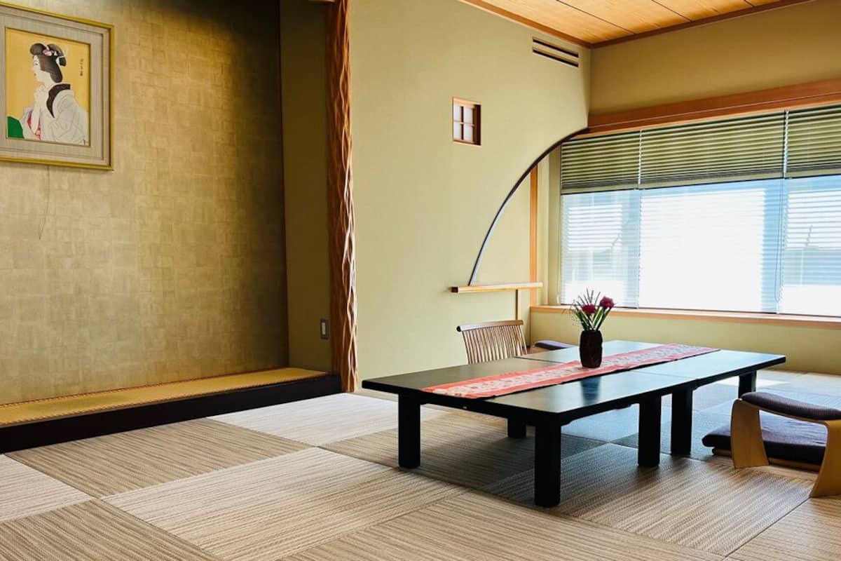 A traditional Japanese room with tatami flooring, a low table, a chair, and a window with blinds.