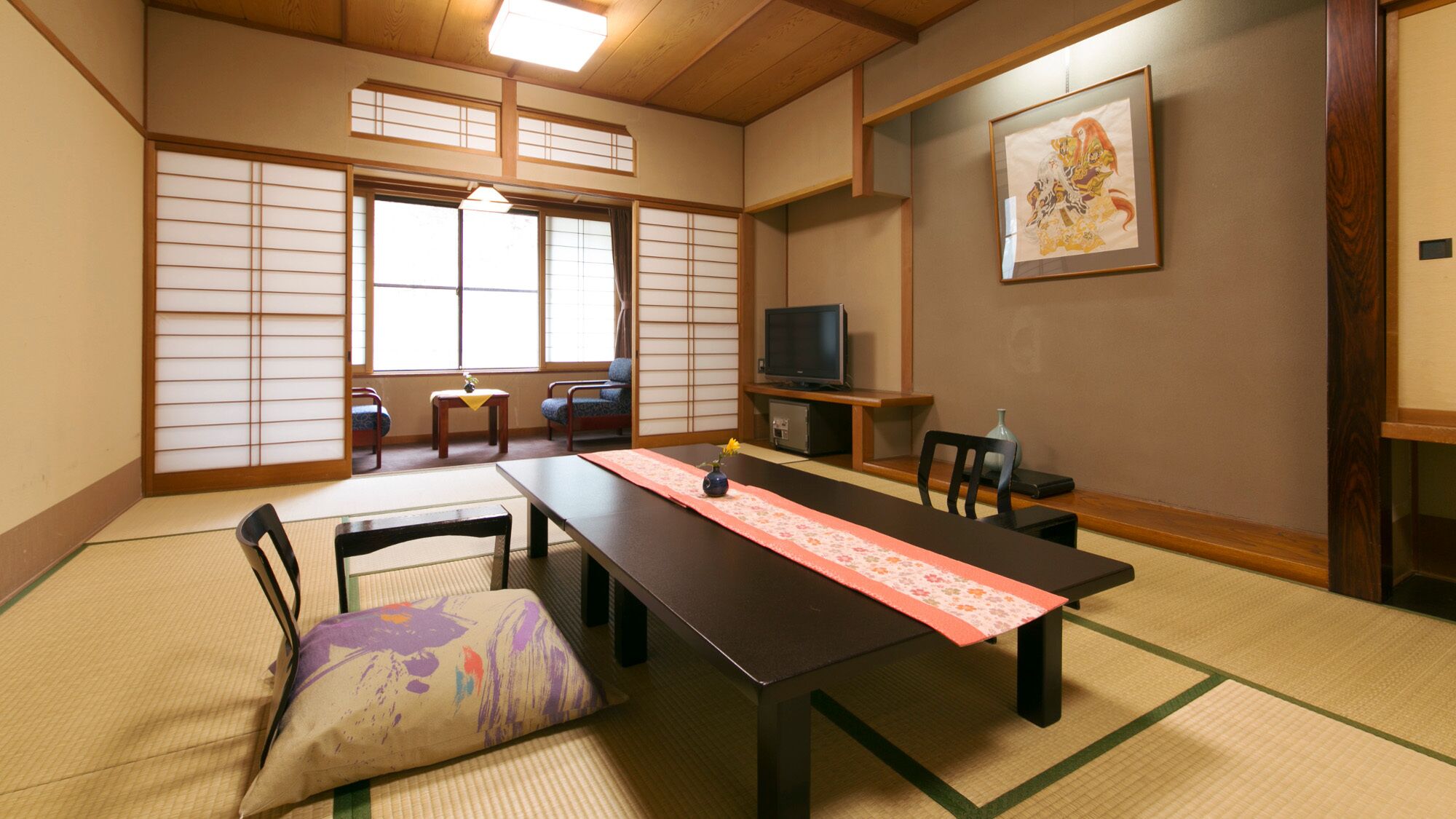 A traditional Japanese room with tatami flooring, a low table with a pink runner, a television, and a framed artwork on the wall.