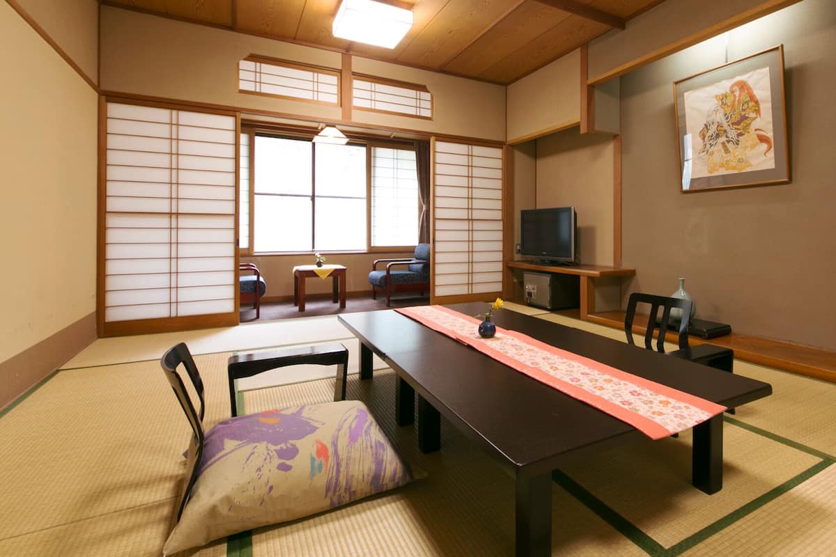 A traditional Japanese room with tatami flooring, a low table with a pink runner, a television, and a framed artwork on the wall.