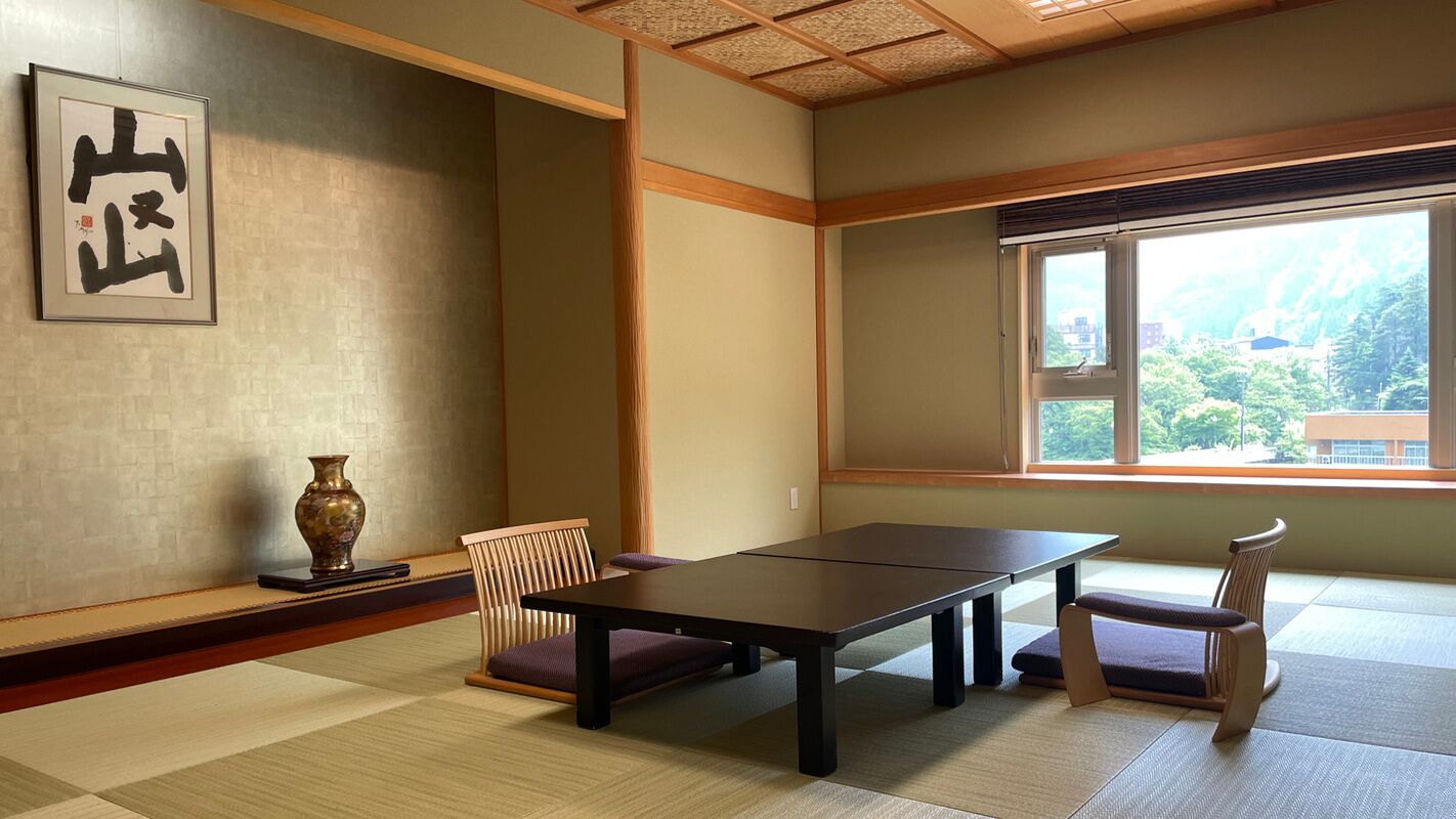 Una habitación tradicional japonesa con suelo de tatami, una mesa baja, dos sillas, un jarrón y una obra de caligrafía enmarcada.