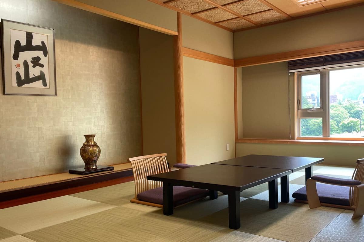 A traditional Japanese room with tatami flooring, a low table, two chairs, a vase, and a framed calligraphy piece.