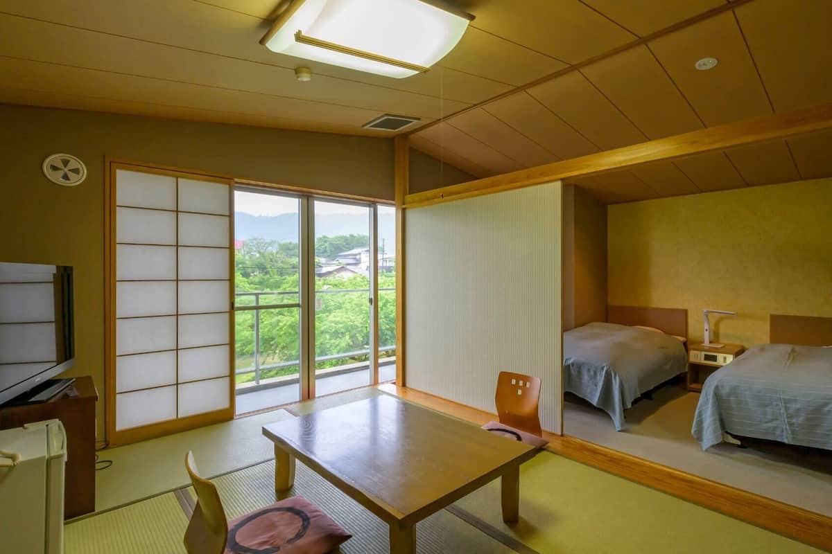 A traditional Japanese-style room with tatami flooring, a sliding shoji screen, a wooden table, two beds, a television, and a view of greenery outside.