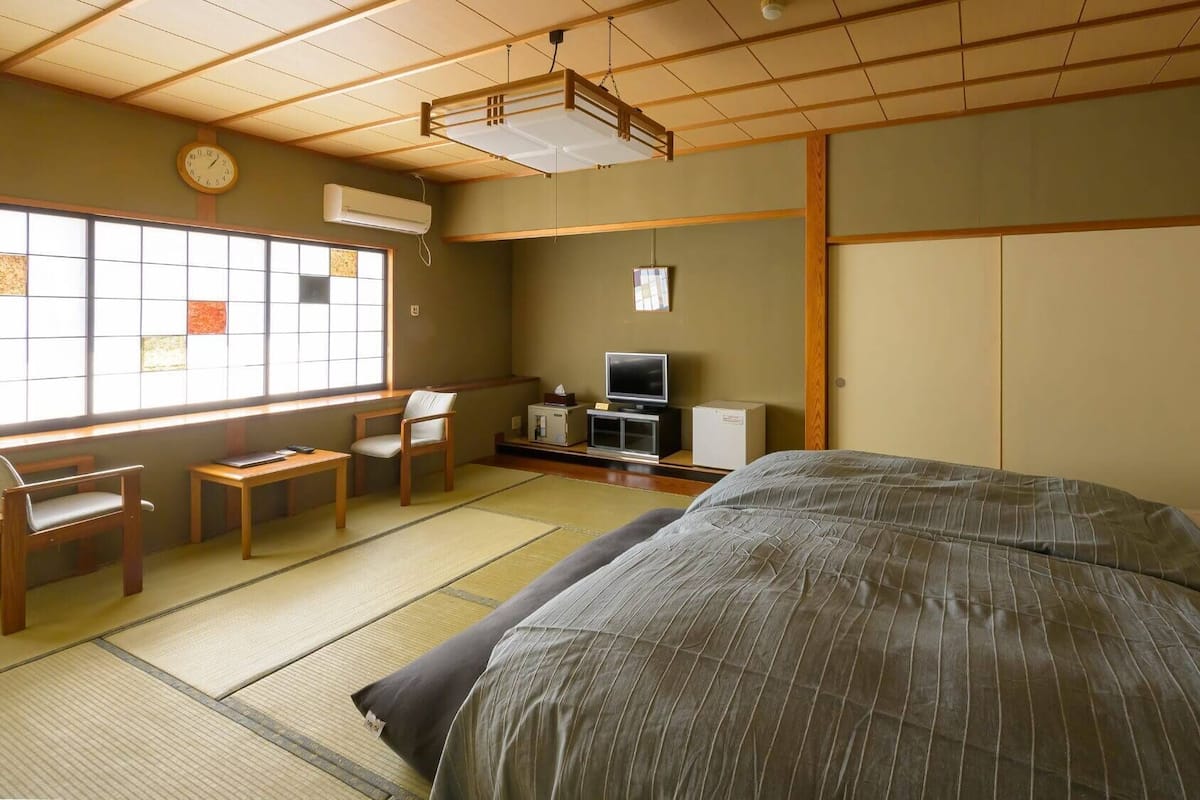 A traditional Japanese room with tatami flooring, a bed, a desk with a chair, a television, and a wardrobe.