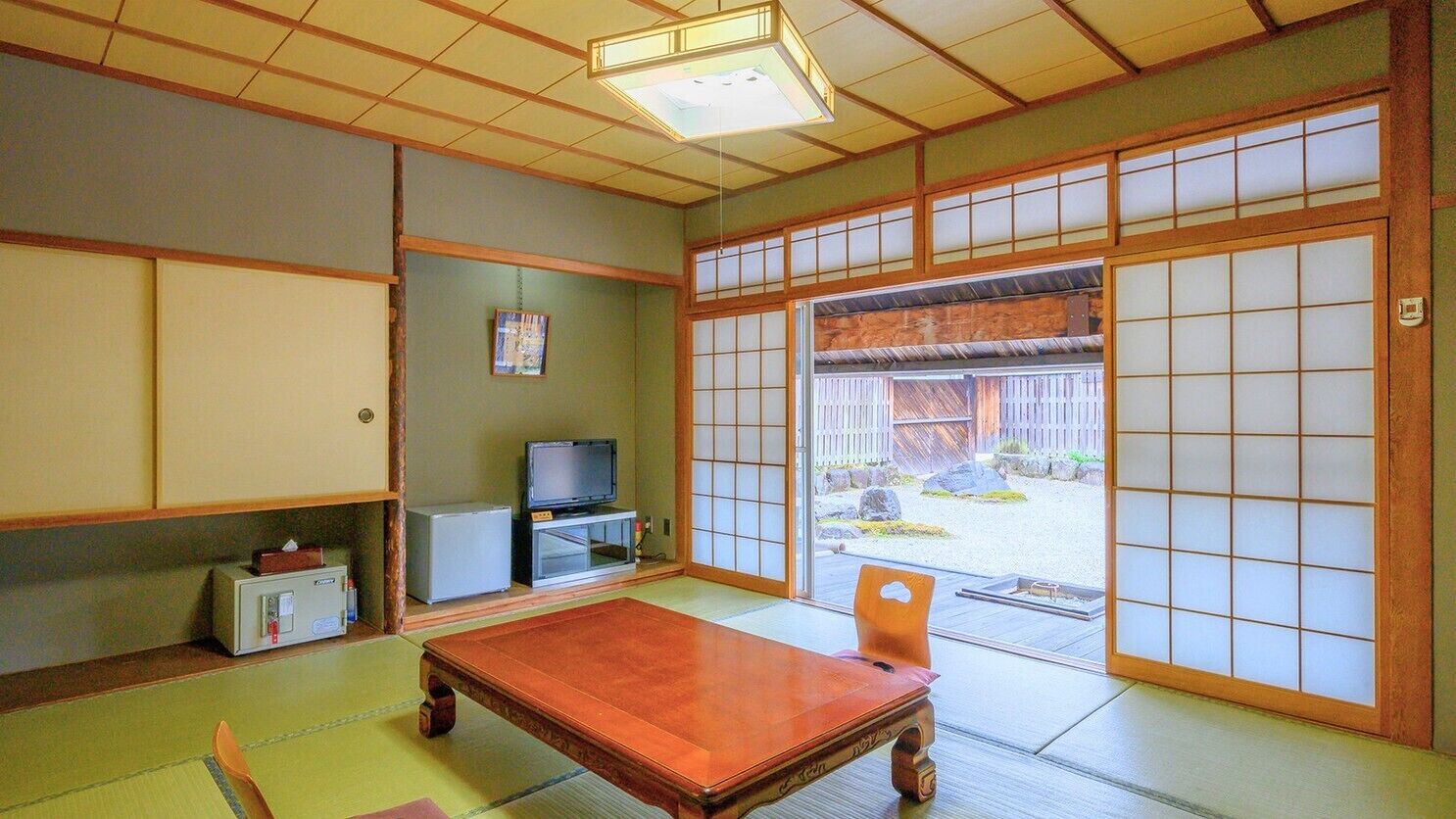Una habitación japonesa tradicional con suelo de tatami, una mesa de madera baja, un televisor y puertas correderas que dan a un espacio exterior.