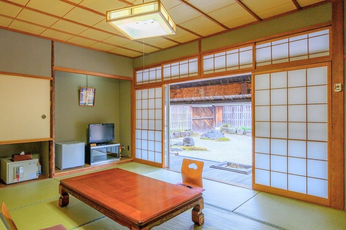 A traditional Japanese room with tatami flooring, a low wooden table, a television, and sliding doors leading to an outdoor area.