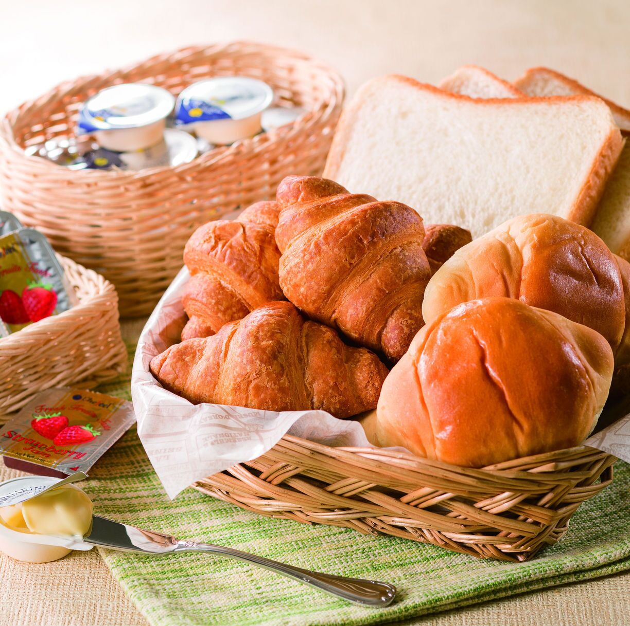 Una cesta de mimbre con croissants y pan, una hogaza de pan blanco, y un tarro de mermelada.
