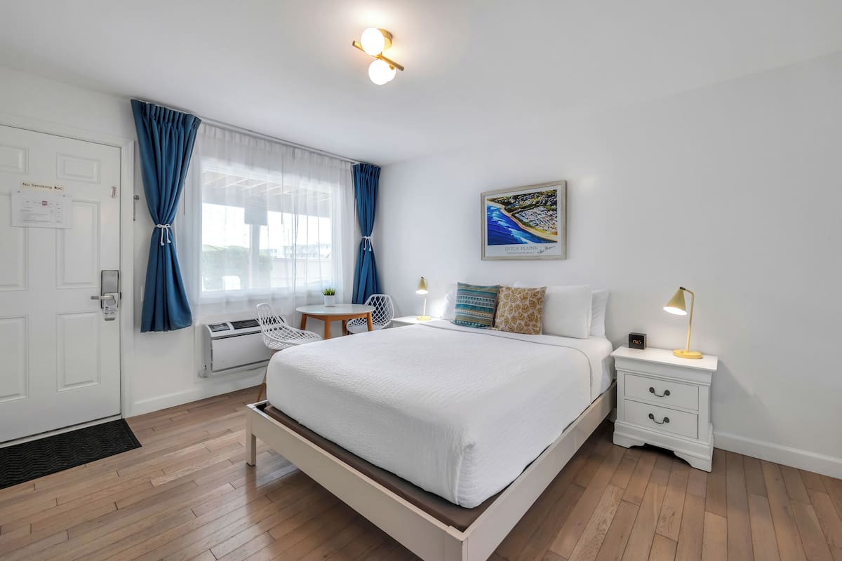 A bedroom with a bed, bedside tables, a nightstand, a window with curtains, a wall-mounted picture, and a door with a 'No Smoking' sign.