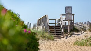 Un portón de madera en la playa con indicaciones, un sendero arenoso y un cielo azul despejado.