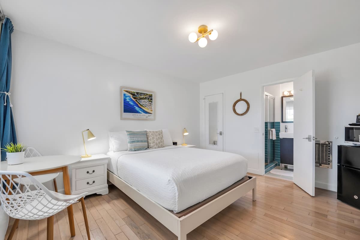A bedroom with a bed, bedside tables, a desk, a chair, a wall-mounted lamp, and a door leading to a bathroom.