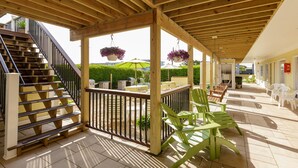 Un espacio al aire libre techado con una escalera de madera, sillas verdes y cestas de flores colgantes.