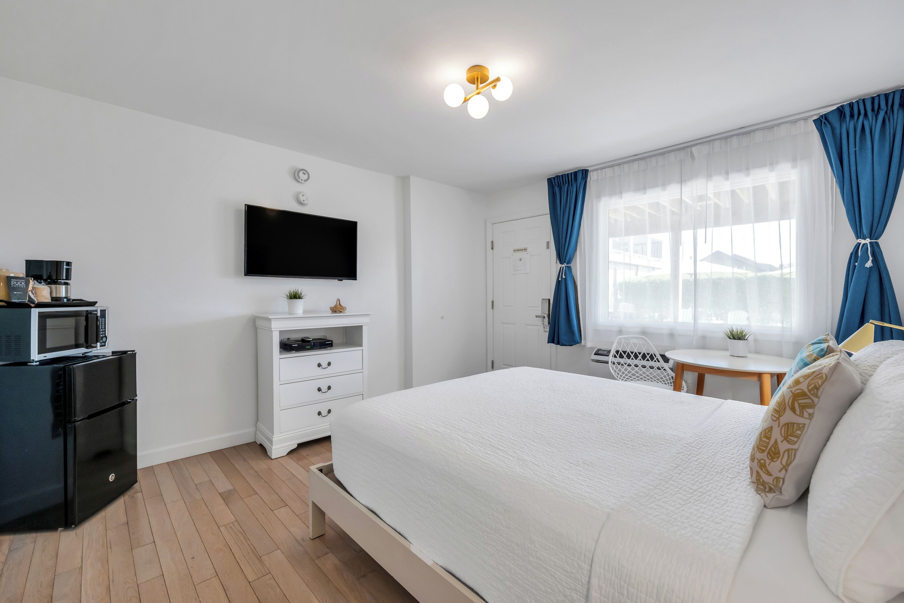 Un dormitorio con cama, televisión montada en la pared, una mesita con silla, microondas y refrigerador.