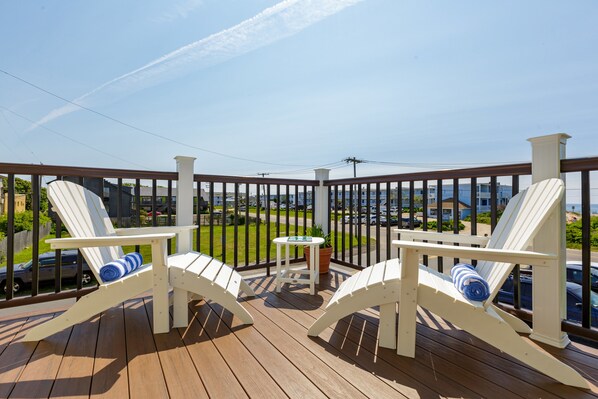 A wooden deck with two white Adirondack chairs and a small table, overlooking a parking area and buildings.