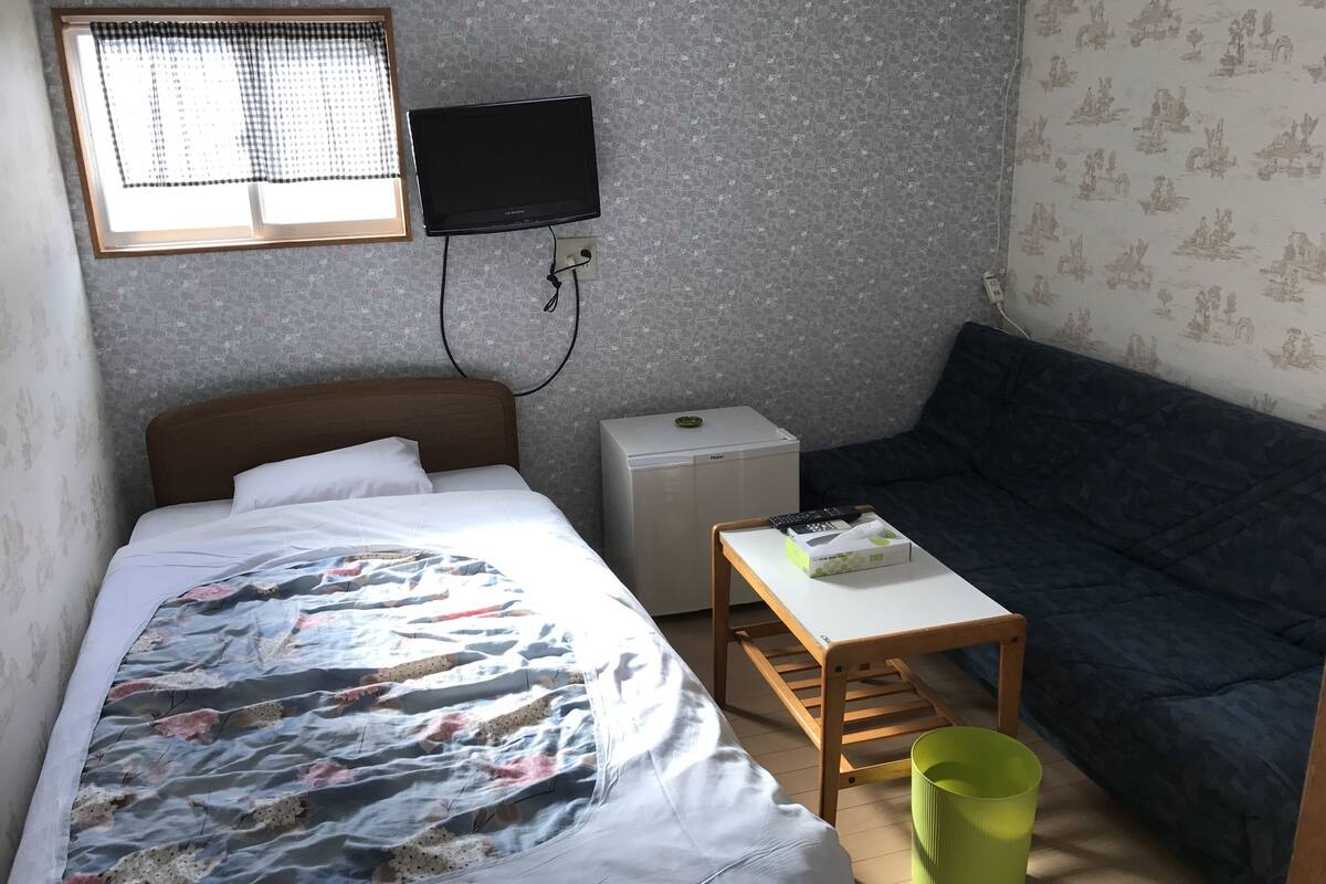 A small, neatly arranged room with a bed, a sofa, a small table, and a television mounted on the wall.