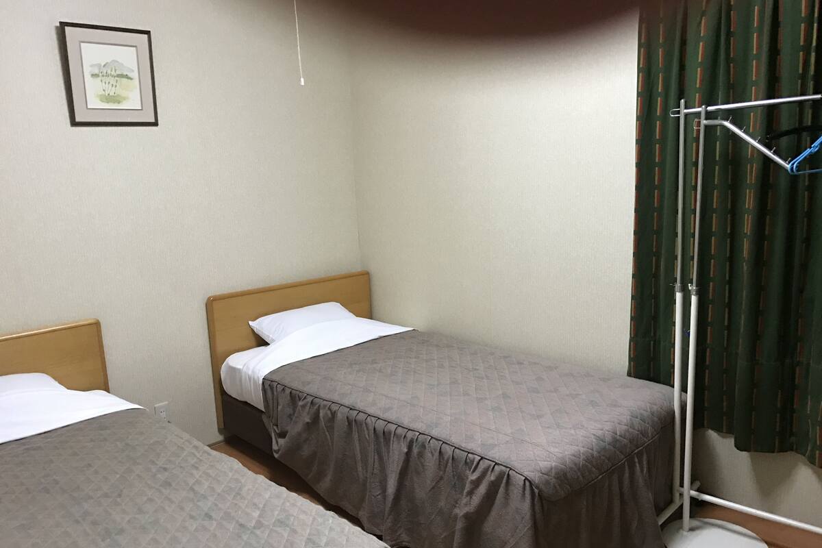 A small, clean room with two single beds, a bedside table, a hanging light, and a curtain.