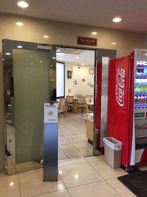 Entrada de hotel com máquina de refrigerantes Coca-Cola, placa indicando sala de chá e sinalização de serviço de café.