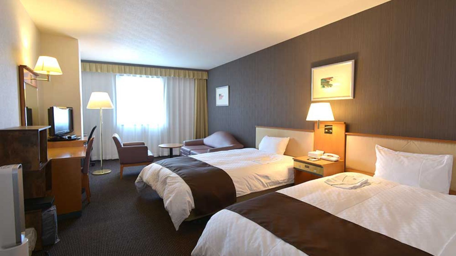 Habitación de hotel con dos camas, un escritorio con computadora, una silla, una mesita y un ventanal con cortinas.