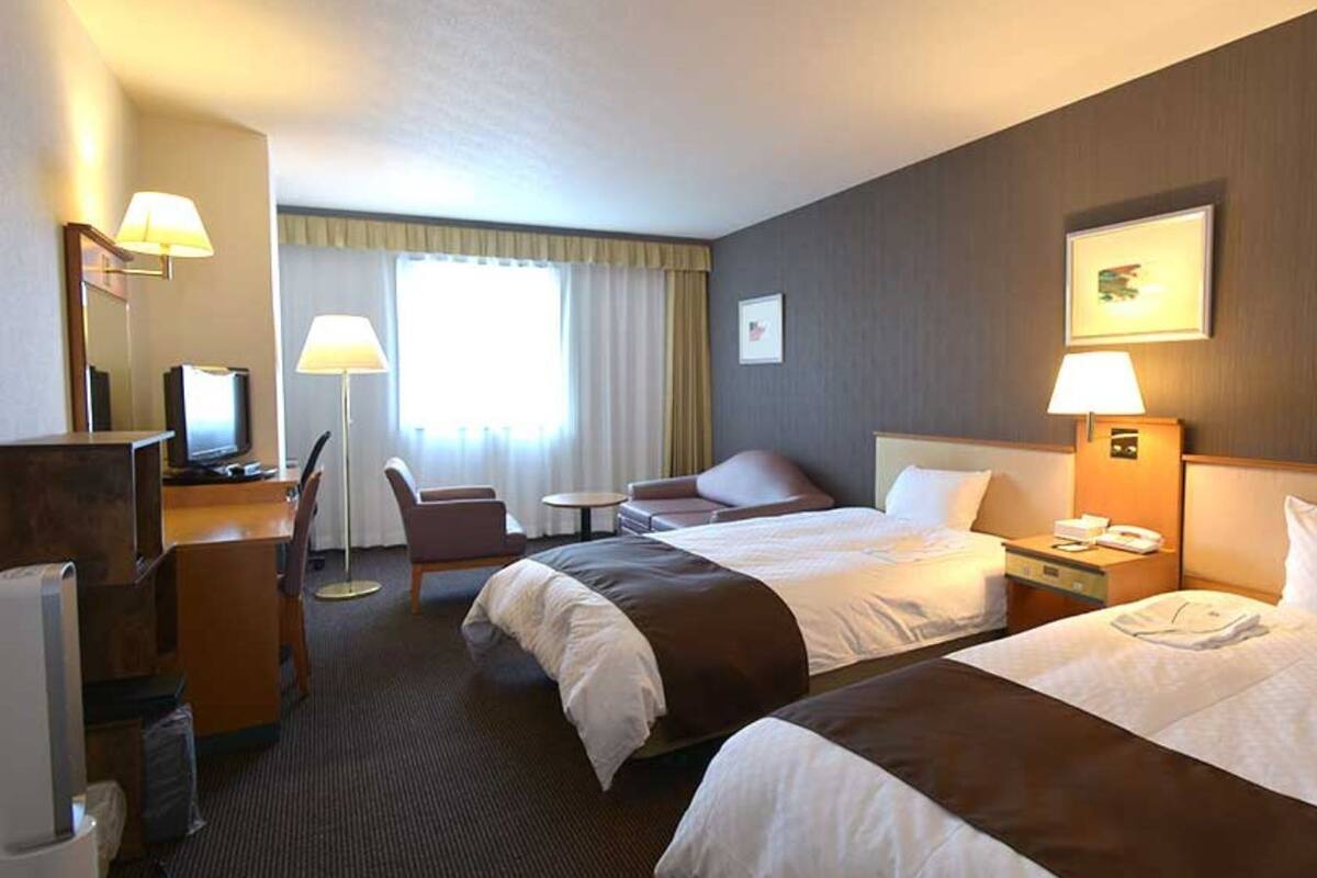Habitación de hotel con dos camas, un escritorio con computadora, una silla, una mesita y un ventanal con cortinas.