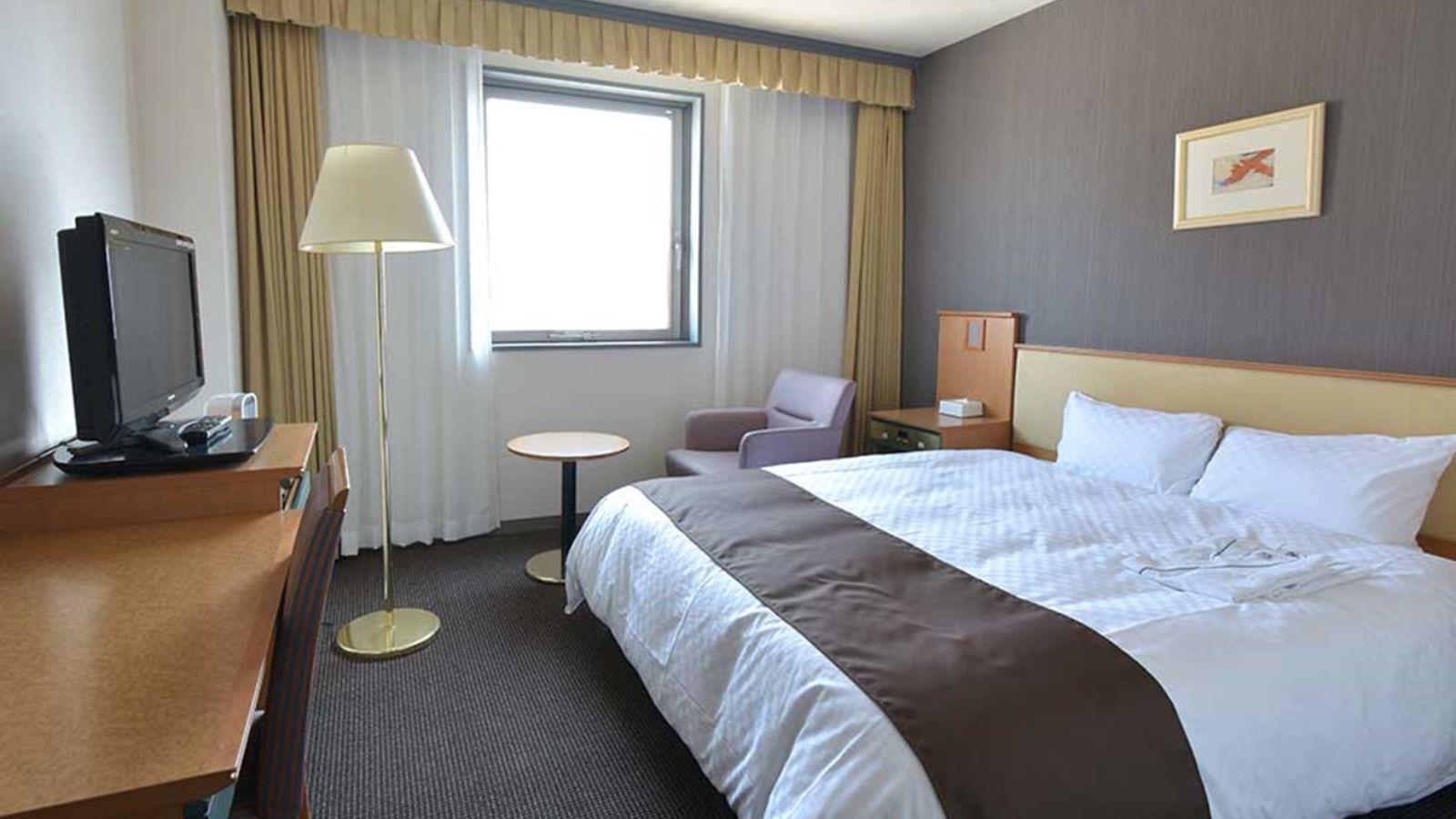 Habitación de hotel con cama, escritorio con televisor, silla, mesita y ventana con cortinas.