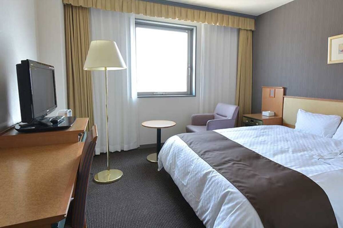 Habitación de hotel con cama, escritorio con televisor, silla, mesita y ventana con cortinas.