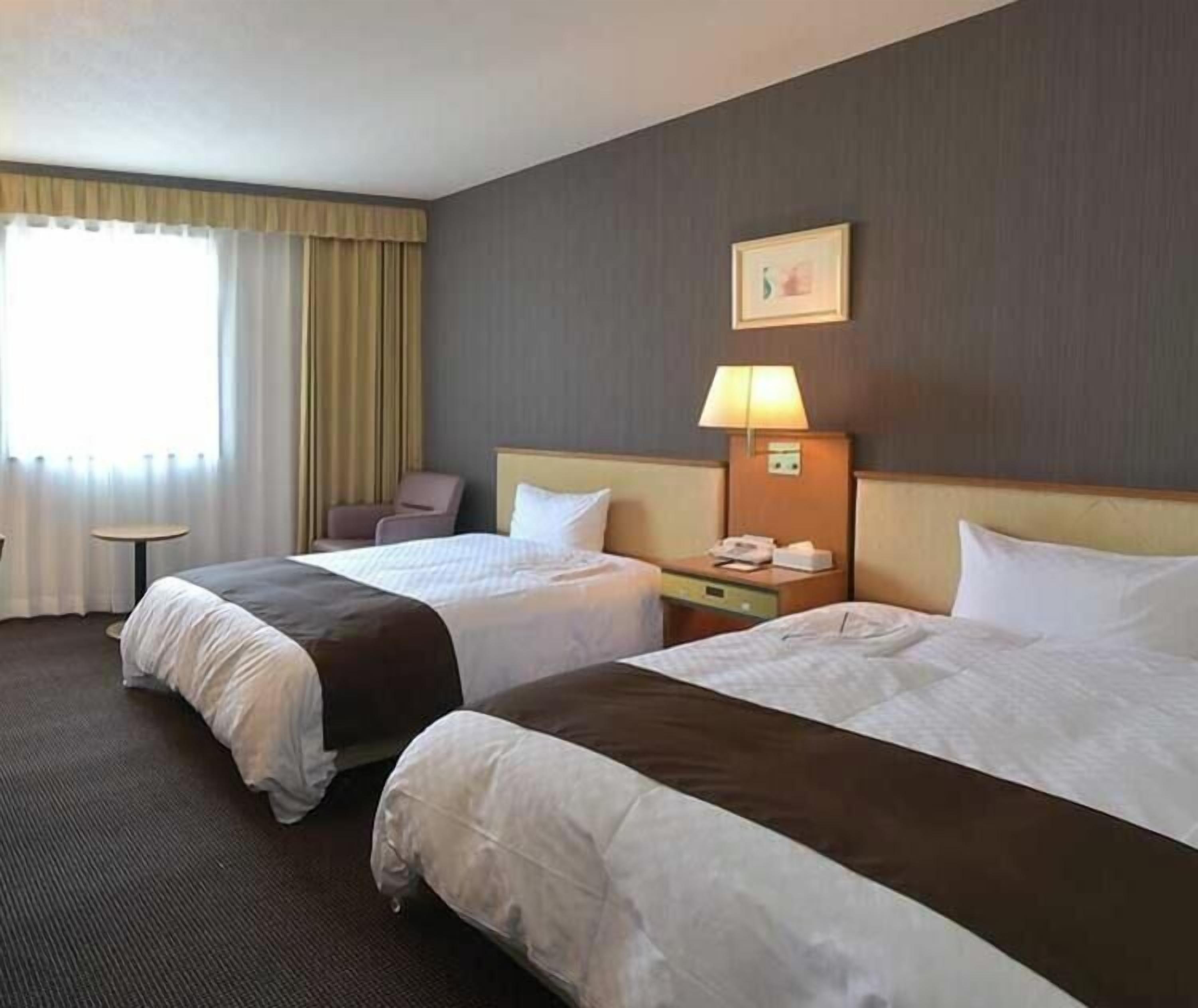 Habitación de hotel con dos camas, una mesita, una silla, una lámpara y un cuadro en la pared.