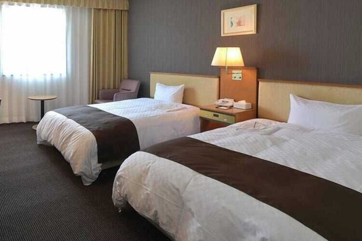 Habitación de hotel con dos camas, una mesita, una silla, una lámpara y un cuadro en la pared.