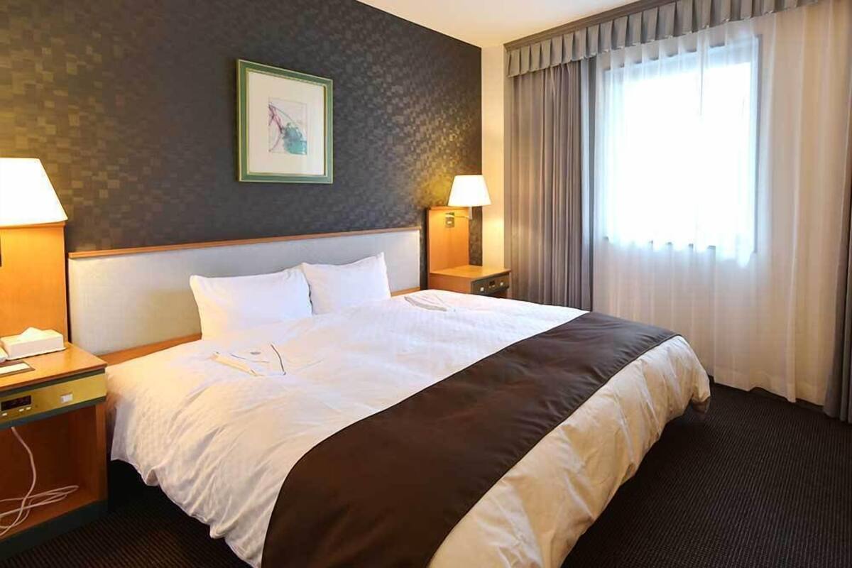 Habitación de hotel con una cama grande, dos lámparas de noche, un cuadro enmarcado en la pared y una ventana con cortinas.